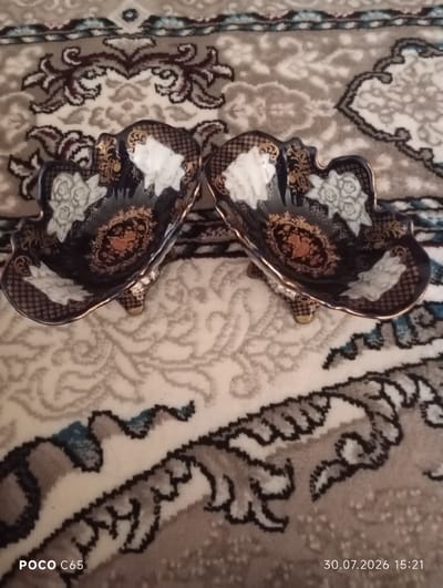 The image displays two decorative, dark blue porcelain bowls with gold and white ornate detailing, placed on a patterned carpet. The bowls are shaped with a fluted edge and have raised floral motifs in white and gold. Each bowl rests on three small, gold-tipped feet. The carpet has a complex, repeating pattern in shades of beige, brown, and white, with black outlines. The lighting suggests an indoor setting, likely a domestic environment. The watermark "POCO C65" is visible in the lower left corner, and the date "30.07.2026 15:21" is in the lower right corner. There are no people present, and no specific location in the city of Beruniy, Uzbekistan is indicated.