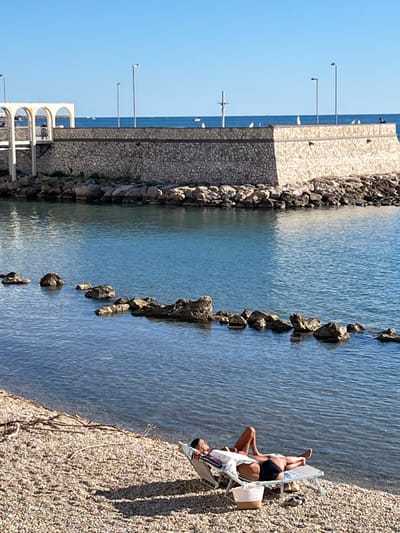 The image displays a sunny coastal scene in Civitavecchia, Italy, featuring a person relaxing on a pebble beach in the foreground.

In the lower right, a female individual with short dark hair lies supine on a light-colored reclining beach chair. She wears dark bottoms and a light-colored open shirt over a lighter top, with her arms crossed over her stomach and knees bent. A light-colored woven tote bag with brown straps rests on the gravel next to the chair, and a magazine or book is visible on the chair beneath her arm. The beach consists of small gray and tan pebbles, with some driftwood branches visible on the left.

The midground features calm, blue-green water. A line of natural rocks partially submerged and breaking the surface extends from the mid-left towards the lower center. Beyond the water, a robust seawall or pier constructed from rough-hewn stone forms a barrier, topped with a lighter, smooth concrete cap. Multiple tall streetlights are evenly spaced along the top of this wall. On the far left, an elevated white structure with several arches is visible, and two indistinct figures are present on its walkway. To the right of the arched structure, a white pole with a horizontal crossbar (possibly a navigational aid) stands on the seawall. On the far right end of the seawall, a small, dark figure is seated.

The background shows a clear, bright blue sky meeting the open sea at the horizon. Scattered across the distant water are numerous small white sailboats and one small dark motorboat.
