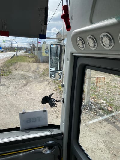 This image captures the interior view from a vehicle, likely a bus, looking out through the front and side windows. The perspective is from the driver's area, facing forward and slightly to the right.

Inside the vehicle, three circular air vents with metallic finishes are visible on the ceiling panel above. A small red, plush object, possibly a toy or decoration, hangs from the ceiling near the rearview mirror. The rearview mirror itself reflects part of the vehicle's interior, showing seats or interior structures. A black device, resembling a microphone or sensor, is attached to the windshield near the top right. A translucent sign or document is affixed to the passenger side of the windshield with suction cups and black binder clips, displaying partially visible Cyrillic text, "ПОР" (likely part of a word like "ПОРЯДОК" meaning "order" or "procedure"). Another grey fabric panel, possibly a sunshade or curtain, is held by binder clips on the left side of the windshield frame. On the vehicle's dashboard or side panel, the brand name "YUTONG" is visible.

Outside the vehicle, the scene reveals an unpaved, dusty ground area with scattered debris like rocks and broken concrete, along with patches of sparse grass. In the distance through the top window, a rusty metal box is mounted on a pole near what appears to be a concrete barrier. Through the main windshield, a road with a crosswalk sign and a main road sign (blue square with a yellow arrow) is visible further away. Utility poles and wires crisscross the sky. Several small flags, one clearly the Russian flag and another blue and white, are visible hanging from a structure in the mid-ground, alongside a banner depicting the Earth from space on a yellow background. The sky is bright and clear, indicating daytime and good weather.

The scene suggests the vehicle is either stationary or moving slowly through a semi-rural or industrial area in Bol'shoe Teshkovo, Russia.