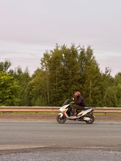 A person wearing a dark jacket and a red helmet is on a scooter on the side of a road. The scooter is white and black. The person is leaning over the handlebars, appearing to be either adjusting something or preparing to move. The scooter has its kickstand down, supporting it. In the background, there are many green trees, suggesting a rural or suburban setting with a lot of vegetation. The road is paved and grey, with a concrete barrier on the right side. The sky is overcast and light, indicating it might be daytime, possibly morning or late afternoon, with a possibility of rain or a cloudy day. There are no visible text elements in the image. The overall impression is a pause in a journey, with the rider and their scooter on the edge of the road, surrounded by nature.