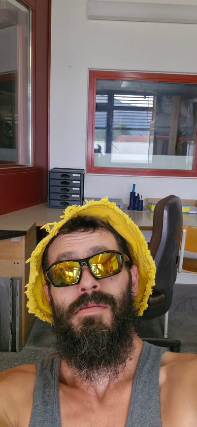 A close-up, eye-level, selfie-style photograph features a man with a full beard and a yellow bucket hat with frayed edges. He is wearing reflective sunglasses with orange accents and a grey tank top. The background appears to be an indoor workspace or office, likely in Uetendorf, Switzerland, given the context provided. Elements visible include a large window with red framing, a filing cabinet, a desk, a rolling chair, and office supplies. The lighting suggests an indoor setting with natural light contributing to the overall illumination.