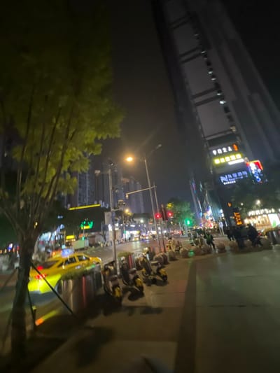 A nighttime urban scene features a street lined with parked motorcycles. A yellow taxi cab is visible in the lower right, suggesting traffic. Buildings with lit-up signs and streetlights create a bright city atmosphere. Green trees are seen in the lower left corner. The photograph appears to be taken from a slightly elevated position, adding depth to the urban landscape.