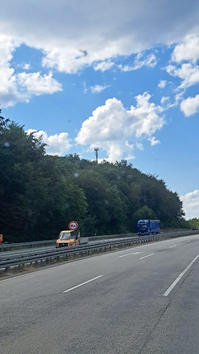 This is a vertical, eye-level, outdoor, daylight, full-color photograph taken from a vehicle driving on a highway. The highway is paved with asphalt and marked with white lines. In the foreground, the road stretches forward, with a lane marker to the right and a merging arrow to the left. A metal guardrail separates the highway lanes from the adjacent embankment. The embankment is densely covered with green trees, forming a thick forest that extends to the top of the frame. A tall, slender telecommunications tower is visible above the trees on the ridge.
On the highway, in the middle ground, a small, orange utility truck with a flatbed is traveling in the left lane. It has a circular traffic sign mounted on its roof, which appears to be a prohibition sign, though the specific symbol is not clearly discernible. To the right of the utility truck, a blue semi-trailer truck is in the right lane, also traveling forward. The sky above is a bright blue with scattered white cumulus clouds, some of which are large and puffy. The overall impression is of a sunny day on a highway bordered by a dense forest. This scene is consistent with a location within the administrative boundaries of Siegen, Germany, given the highway infrastructure and surrounding natural landscape. No individuals are clearly visible in the photograph.