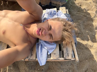 This media file shows a young person lying on a wooden surface, possibly a beach chair or pallet, on a sandy and pebbly area. The person is lying on their back, facing the camera with their head tilted slightly to the side. They have blond hair that is messy and windswept. A light blue towel is draped over their chest and head.
The person's face is contorted into a playful or silly expression, with their eyes squinted and their mouth open, sticking out their tongue slightly. They are wearing a purple cord necklace with a small, silver cross pendant. The sunlight casts shadows on the sand, suggesting it is daytime. The overall impression is one of leisure and fun on a sunny day, possibly at the beach or a similar outdoor location in Gres-2, Kazakhstan. No visible text is present in the media file.