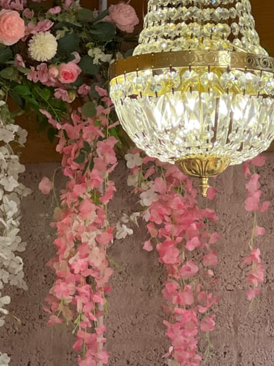 The image shows a close-up view of a crystal chandelier hanging amidst artificial floral decorations. The chandelier has a brass-colored frame and is adorned with numerous clear crystal prisms that reflect light. Cascading down from the upper left and right are garlands of pink and white artificial flowers, resembling wisteria or cherry blossoms. The background appears to be a textured wall, possibly stone or stucco, in a muted brown or grey tone. The lighting is warm, highlighting the sparkle of the crystals and the soft hues of the flowers. The scene is devoid of any people or animals. There is no indication of the location being in Donegal, Ireland, as the image provides no geographical or identifying markers for that specific place.