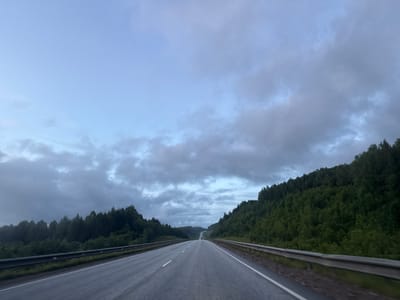 The media file captures a scenic view of a highway in Div'ya, Russia, likely during the late afternoon or early evening, judging by the soft, diffused light and the overcast sky. The road, paved and in good condition, stretches into the distance, disappearing over a gentle incline. Guardrails line both sides of the asphalt, their metallic sheen reflecting the muted light.
The surrounding landscape is dominated by lush, dense evergreen forests that climb the rolling hills. The trees are a vibrant green, creating a stark contrast with the grey, cloudy sky. Wisps of clouds drift across the sky, obscuring the sun and casting a slightly melancholic or peaceful mood over the scene. The overall atmosphere is one of serene travel through a natural, undeveloped area. There are no visible people or text in the image.