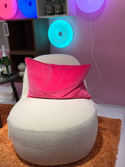 The image depicts an interior scene featuring a round, textured cream-colored armchair positioned on a fluffy orange rug. A bright pink velvet pillow rests on the armchair. In the background, a wooden shelf unit holds decorative items and bottles. Several circular, illuminated wall lights in pink, blue, and teal are visible against a pink wall, suggesting a modern and vibrant decor. The scene is situated in the city of Son en Breugel, Netherlands. There are no people visible in the image.