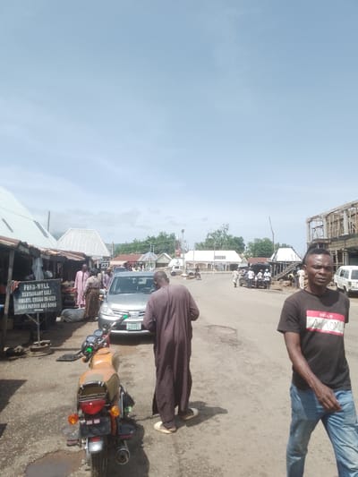 This media file depicts a bustling street scene in Beli, Nigeria. The image features several individuals, including a man in the foreground wearing a black t-shirt with the text "HELLO AWESOME" and blue jeans, walking towards the viewer. To his left, another person in a brown robe walks away from the camera. In the background, a silver sedan is parked, and several other people are visible near shops and along the street. A motorcycle is parked in the lower left quadrant of the image.
The setting appears to be a market or commercial area, with various structures that resemble shops or stalls lining the street. One stall has a sign that reads "FEED-WELL RESTAURANT BALI NWANYI, ENUGU ISI-UZO MAMA FAVOUR AN SONS." The road is unpaved and appears somewhat worn. In the distance, more buildings and trees are visible, suggesting an urban or semi-urban environment. The sky is bright and clear, indicating it is daytime with fair weather. The overall impression is one of everyday activity and commerce. The presence of multiple people and vehicles suggests a busy period.