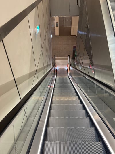 The image shows two parallel escalators located at the Thiais RER station in Thiais, France (latitude: 48.747, longitude: 2.373). The escalators are empty, and the camera is positioned at the top of the escalators, looking down. The escalators are made of metal and glass. The walls of the station are made of concrete and metal. There are no people present in the image. There is a single light fixture on the wall to the left. The top of the escalators is illuminated with overhead fluorescent lights. There is a doorway at the top of the escalators, leading to an unknown area. The escalators are both ascending.