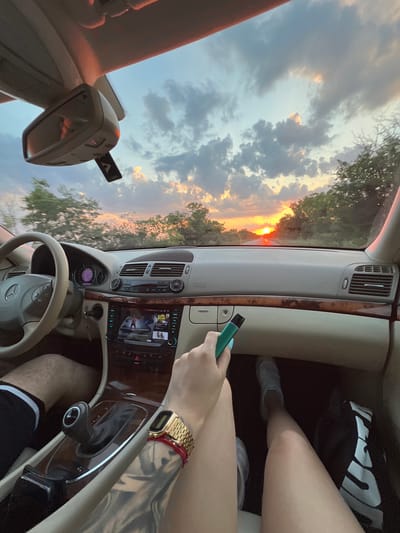 The image shows the interior of a car at sunset. The sun is low on the horizon, casting a warm glow through the windshield and illuminating the clouds. Trees and foliage are visible outside the car, suggesting a rural or suburban setting.
Inside the car, a person's hand holds a green vaping device. Another person is visible in the passenger seat, with their legs and feet showing. They are wearing shorts and sneakers. A gold watch is worn on the wrist of the person holding the vape. The car's dashboard and center console are visible, with a navigation screen displaying what appears to be a music interface with two women's faces. The car's interior is light-colored, with wood trim accents.
The time of day is sunset, and the weather appears to be clear with scattered clouds. The location is identified as Velichkovo, Bulgaria. The overall mood conveyed is relaxed and casual. There is no visible text other than what may be on the car's display screen.