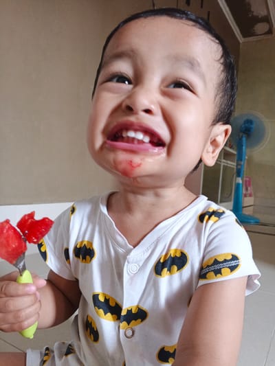 A close-up, eye-level shot shows a young child with dark hair, looking up and to the right with a wide, somewhat contorted grin. Their mouth is open, revealing white teeth and a red stain around the lips and chin, suggesting they have been eating something like watermelon. The child is wearing a white t-shirt with a repeating pattern of yellow and black Batman symbols.

In the child's left hand, they hold a green utensil, possibly a fork or spoon, with two pieces of red fruit, likely watermelon, impaled on it. The background is slightly out of focus, showing a beige wall and a blue oscillating fan in the distance. The lighting suggests it is daytime. The child's expression is one of extreme excitement or perhaps mild discomfort, with eyebrows raised and cheeks scrunched. The setting appears to be indoors, possibly in a home environment in Cibungbulang, Indonesia. There is no visible text in the image.