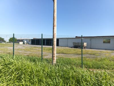 This image captures a wide shot of a fenced industrial area on a clear, sunny day. The foreground is dominated by lush, green grass that appears slightly windblown. A wire mesh fence with green posts runs horizontally across the middle ground, separating the viewer from the industrial scene beyond.
Behind the fence, a large, utilitarian building with a corrugated metal exterior and a visible roofline stretches across the frame. To the left of the building, a few trees are visible, and further to the left, a smaller structure can be seen. A light-colored pole, appearing to be made of wood and showing signs of rust, stands prominently in the mid-ground, directly in front of the large building and partially obscuring it.
In front of the building and behind the fence, there is a dirt or gravel area, with some evidence of patchy vegetation. A light-colored, flatbed trailer is parked near the building on the right side of the image. A sign with a green and yellow graphic is affixed to the exterior of the building on the far right. The sky above is a clear, pale blue. The scene suggests an outdoor, possibly agricultural or light industrial, setting in Tenevo, Bulgaria. There are no people or animals visible in the image.