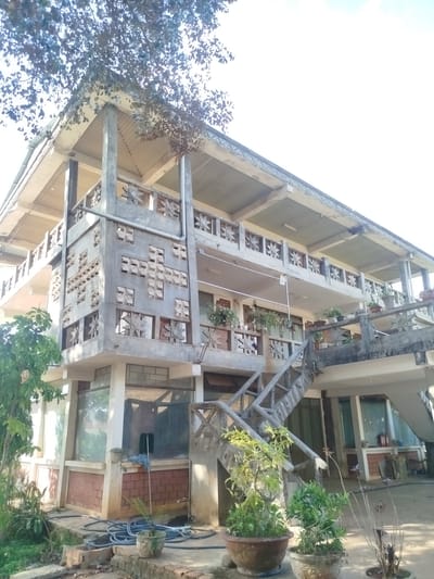 A two-story building stands in An Phu Ward, Vietnam. The building has a unique design with concrete walls featuring geometric cutouts. The lower level has glass windows and brick sections. A concrete staircase leads to the upper level, adorned with potted plants. The building is surrounded by foliage. A coiled blue hose lies on the ground near the base of the building.