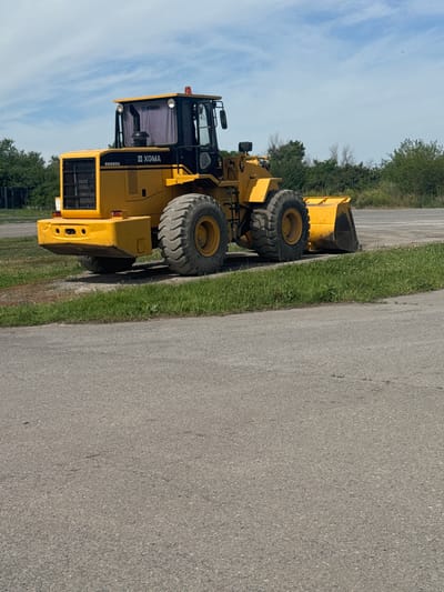 A yellow XGMA wheel loader is parked on an asphalt surface next to a strip of grass. The wheel loader is facing right and is viewed from the front-right. The machine has large, treaded tires, a raised cab with tinted windows, and a bucket at the front. The background features a line of green trees and a cloudy blue sky. The scene appears to be an outdoor industrial or construction area. The location is in Shakhty City District, Russia.
