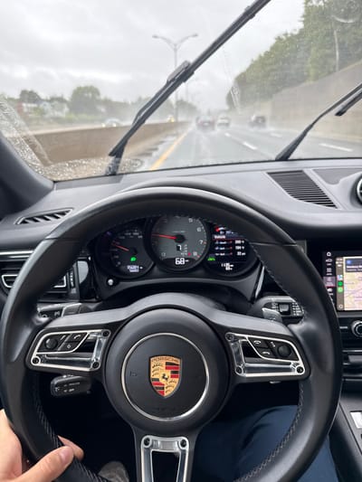 The image depicts the interior of a Porsche vehicle from the driver's perspective, looking out onto a wet highway during rainfall in Newton, United States. The driver's left hand is visible at the bottom left, resting on the leather-wrapped steering wheel, which prominently displays the Porsche crest in its center.
The instrument cluster is visible through the steering wheel, showing a speedometer indicating a speed of 68 (presumably MPH) and a tachometer. Digital readouts include an odometer showing 35004 miles, vehicle information such as 207 miles, 194°F, and 22 psi, and a time of 11:35. The windshield is covered with raindrops, and the wipers are active, with the right wiper extended upwards.
Outside the vehicle, a multi-lane highway is visible with wet asphalt, reflecting ambient light. Several vehicles are ahead in the distance, appearing blurred due to motion or atmospheric conditions. To the left of the highway, residential buildings and trees are partially visible behind a barrier. To the right, a concrete barrier separates the highway from what appears to be more dense foliage. An overhead street light structure spans the highway. The sky is overcast, consistent with rainy weather.