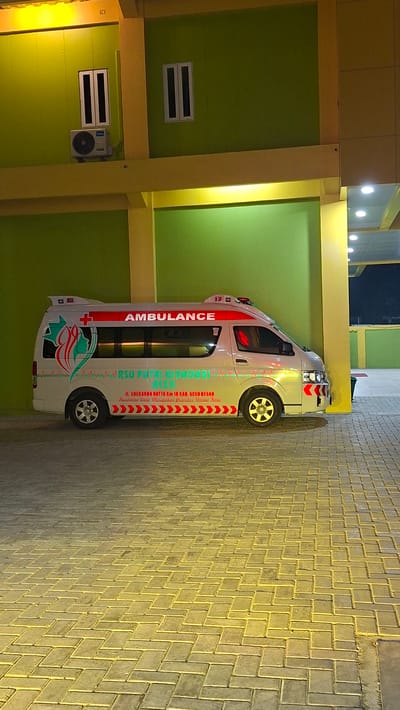 This media file shows an ambulance parked outdoors at night. The ambulance is white with red and green graphics and text. The text on the side of the ambulance reads "AMBULANCE," "RSU PUTRI BIDADARI," "ACEH," and "JL. SOEKARNO HATTA KM 18 KAB. ACEH BESAR" and "Kesehatan Anda Merupakan Prioritas Utama Kami". The ambulance appears to be well-maintained.

The setting is an outdoor area with a green wall on the left and a paved walkway on the right. The walkway is made of rectangular pavers arranged in a herringbone pattern, illuminated by yellow lighting. The green wall has two rectangular vents and an air conditioning unit mounted on it. The overall scene suggests an institutional setting, possibly a hospital or medical facility, given the presence of the ambulance and the green walls. The lighting indicates it is nighttime, and the weather appears to be clear. There are no people visible in the image.