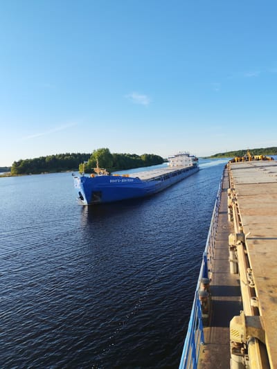 The image shows a wide, dark blue river under a clear blue sky with a few wispy clouds. On the river, a large blue barge with "ВОЛГО-ДОН 5069" written on its side is moving away from the viewer. The barge has a white superstructure at the stern and a long, flat deck. To the right of the frame, another barge, likely a cargo barge, is positioned closer to the viewer. Its deck is made of weathered, yellowish-brown material. The edge of this barge has a blue railing. In the background, a tree-lined shore is visible, with the trees appearing dense and green. The overall scene suggests a peaceful day on a waterway in Poteryaevo, Russia.