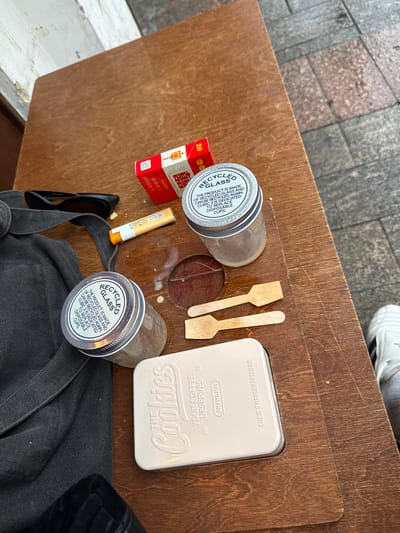 The image depicts a close-up, eye-level view of a wooden table, likely outdoors on a sidewalk, given the cobblestone pattern visible on the right side of the frame. The table is set with several items, suggesting a break or a moment of rest. In the foreground, a light-colored tin box with embossed text that reads "THE COOKIES YOUR COFFEE DESERVES" and "SHANGHAI" is prominent. Next to it are two small wooden stirring sticks.
Behind these, two glass jars with metallic lids are placed on the table. The lid of the jar on the right is clearly visible and features the text "RECYCLED GLASS" and further details about the product's origin and purpose. The jar on the left has a similar lid but is partially obscured by a dark fabric bag.
Further into the frame, a red and white cigarette pack is visible, with Chinese characters on it. To the left of the cigarette pack, a pair of black sunglasses rests on the dark fabric bag. A yellow and orange lip balm or similar stick-shaped item lies between the sunglasses and the cigarette pack. Small splatters of a white substance are scattered on the table surface around the jars. The overall impression is that of a casual setting, possibly a cafe or a street-side table in Xuhui District, China.