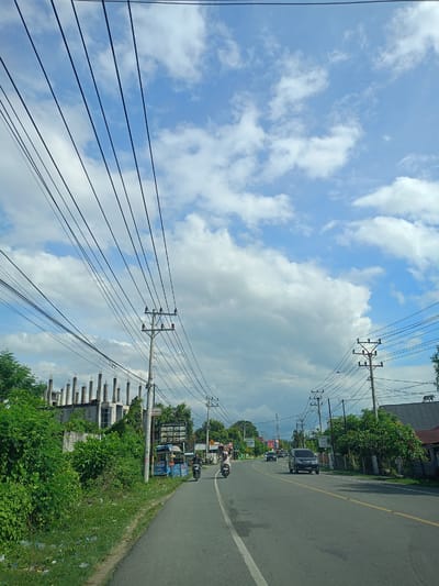 Captured from an elevated perspective, potentially within a moving vehicle, this image presents a street view in Teungoh Baro, Indonesia. The asphalt road, marked by a white lane divider and a yellow side line, leads into the distance, featuring a black SUV and several mopeds. Utility poles, crisscrossed by an array of power lines, line both sides of the road. A building under construction with exposed concrete columns is visible on the left. Lush greenery and vegetation border the road, interspersed with residential houses. A partly cloudy sky occupies the upper half of the frame. A sign for “Modern Cafe” can be seen along the left side of the road.