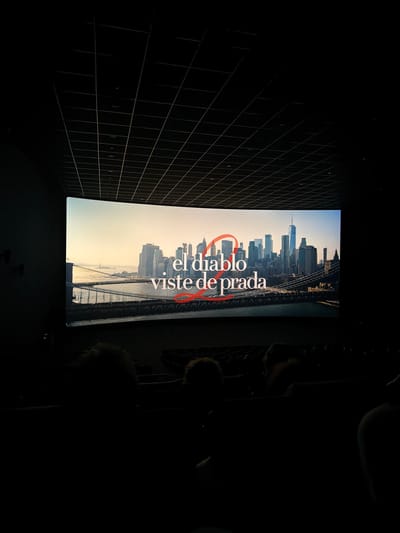 The image captures a movie screening or presentation in a dark room, likely a cinema or auditorium in Paterna, Spain. A large rectangular screen dominates the center, displaying a panoramic cityscape that includes prominent skyscrapers, such as the One World Trade Center, and a suspension bridge, strongly suggesting a view of New York City. The sky in the background of the cityscape is light, indicating daytime or dusk.

Overlaid on the cityscape, bold red text reads "el diablo viste de prada," which translates to "The Devil Wears Prada." A stylized red graphic, possibly resembling a "2" or a heart, is integrated with the text, partially encircling the word "diablo."

The room itself is very dark, with only the bright screen illuminating parts of the foreground. On the left side, a grid-like architectural pattern is visible on the wall, fading into the darkness. To the right and in the foreground, silhouettes of several audience members' heads are discernible, indicating people are seated and watching the screen. The overall atmosphere is that of a typical movie viewing experience.