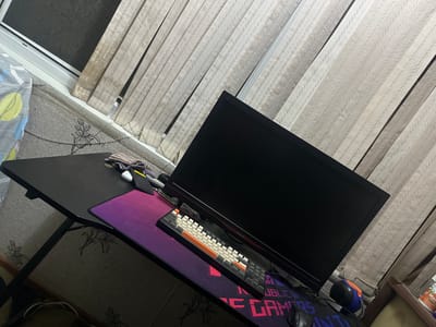 A gaming desk is set up near a window with blinds in a room. On the desk, there is a black computer monitor, a keyboard with white and orange keys, a gaming mousepad with a purple and pink gradient design and "Republic of Gamers" text, a computer mouse, and a small grey object. There is also a black wallet and a small bundle of fabric on the left side of the desk. To the left of the desk, there is a wall with a floral pattern wallpaper. A white and blue patterned blanket is draped over a portion of the desk on the left side. The scene is captured from a high-angle perspective.