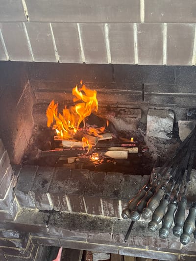 A close-up, overhead shot shows a fireplace filled with burning logs and several metal skewers with wooden handles. The fireplace is constructed of brick, with some of the bricks showing signs of soot and wear. The fire is bright and produces a significant amount of flame and smoke. The skewers are arranged in a row to the right of the fire, with their handles resting on the edge of the fireplace. The setting appears to be outdoors, possibly in a backyard or patio area. The city of Ляды, Russia, is mentioned, suggesting a specific location for this scene.