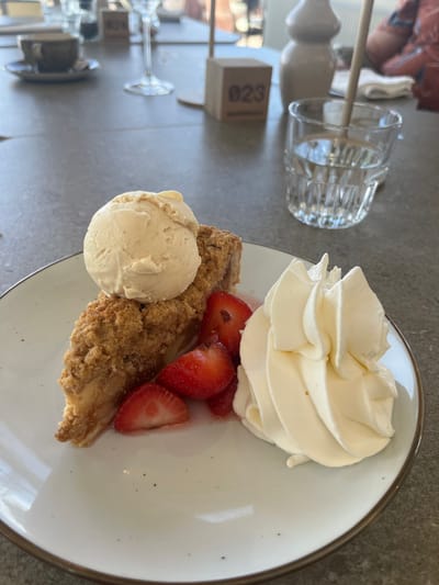 A close-up image captures a dessert served on a light-colored, round plate with a brown rim, positioned on a gray, textured tabletop. The dessert consists of a wedge-shaped slice of apple crumble with a golden-brown, granular topping. A spherical scoop of vanilla ice cream rests on top of the crumble. To the right of the crumble, several red strawberry slices, both halves and quarters, are arranged, with some residual red liquid pooling on the plate. Adjacent to the strawberries and crumble, a generous swirl of creamy white whipped cream is present.
In the blurred background, other table settings are visible on the same gray surface. To the left, a dark-colored cup sits on a saucer, and a tall, empty wine glass stands. Towards the center-right, a light-brown wooden block with the number "023" printed in dark characters is present, serving as a table marker. Further right, a clear glass filled with water, containing a light-colored stirring stick, is visible. Behind the water glass, a light-beige vase or bottle stands. A portion of a person's upper arm or shoulder, clad in a patterned fabric, is partially visible in the upper right background. The scene suggests an indoor dining establishment, specifically inferred to be Anne&Max Overveen, located in Overveen, Netherlands, based on the distinctive wooden table marker style and typical menu offerings.