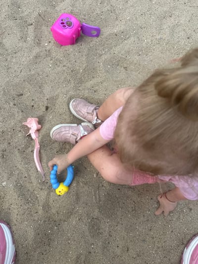 This is a high-angle shot of a young child playing in the sand, likely in Donchovci, Bulgaria. The child is wearing a pink t-shirt and shorts, and light pink shoes with velcro straps. Their hands and legs are dusted with sand.

Scattered around the child are several toys. A bright pink plastic shovel with a purple handle is to the left. A blue and yellow caterpillar-shaped rattle is being held by the child's hand. Another pink toy, possibly a part of a toy set, is lying to the lower right.

The sand appears dry, suggesting fair weather and possibly a daytime setting, though the specific time of day and weather conditions are not explicitly discernible. The overall mood is one of childhood play and exploration. There is no visible text in the image. The child's focus is on the toys and the sand, indicative of engaged play.