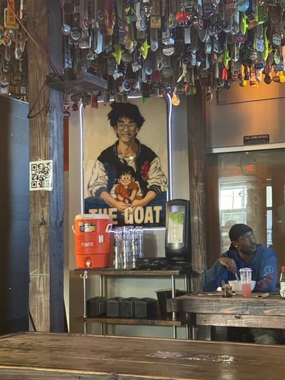 The image presents an interior view of a bar in Spring, Texas, United States. There's a dense array of beer taps to the left, displaying various brand names. A poster with an anime character holding a child is positioned against the wall. Nearby are a drink cooler and glasses. A man sits at the wooden bar counter, interacting with what appears to be a beverage or meal. A "No Smoking" sign is affixed to a light-colored wall or partition. A QR code is visible on a wooden structure.