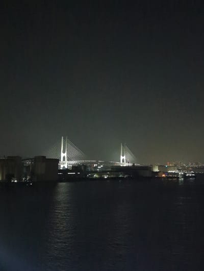 The image shows a nighttime view of the Port of Kobe, Japan, featuring the illuminated Kobe Port Tower and the Rainbow Bridge in the distance. The foreground is dominated by dark water, reflecting the ambient light from the city. In the middle ground, silhouetted structures of the port area are visible, including warehouses and what appear to be ships or ferries. The Kobe Port Tower, a prominent landmark, is illuminated with white lights, and the Rainbow Bridge is also clearly lit. The sky is dark and overcast, common for a night scene in a coastal city. No people are visible in the frame.