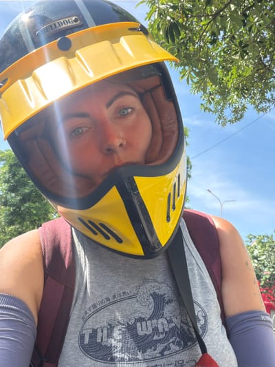 A woman wearing a yellow and black motorcycle helmet, a gray tank top with Japanese text and a wave graphic, and arm sleeves is taking a selfie. She has blue eyes and a nose ring. Behind her, green trees and a blue sky are visible. A streetlamp can be seen in the distance. The scene appears to be outdoors in a tropical environment, likely in the city of Đồng Hới Ward, Vietnam, given the lush vegetation and bright sunlight.