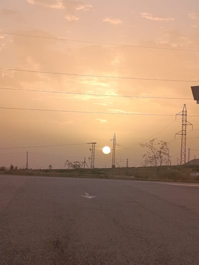 The image depicts a roadside scene with a sunset in the background. A paved road with a white arrow marking dominates the foreground. In the middle ground, a fence runs horizontally, with some sparse vegetation and trees behind it. Several power lines stretch across the frame, supported by tall metal towers. The sun, a bright orb, is visible low in the sky, partially obscured by clouds. The sky is a warm orange and yellow, indicative of sunset. The scene appears to be in an open, possibly semi-rural area, with the characteristic architecture of utility infrastructure suggesting a location near human settlements. Based on the visual cues, this scene could be located in the city of Santomera, Spain, given its landscape often includes such power lines and open terrains. There are no people or animals visible in the image.