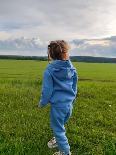 A young child stands in a grassy field with a broad expanse of green land extending towards a line of trees in the distance. The sky is partly cloudy with patches of light breaking through. The child, viewed from behind, wears a blue hooded tracksuit and light-colored shoes. The child has braided hair and is oriented towards the open landscape. The location is Горняк, Russia.