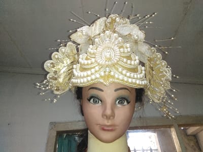 The image displays a mannequin's head wearing an elaborate, ornate headdress, positioned indoors. The headdress is primarily cream, white, and gold, extensively adorned with faux pearls of various sizes, intricate beadwork, and shimmering fabric elements resembling leaves and flowers. Silver-colored wires tipped with small pearls protrude outwards, creating a halo effect. The central part of the headdress features a crown-like structure with multiple tiers of pearls and a large floral motif. The mannequin has a light brown skin tone, dark, stylized eyebrows, large wide-open eyes with light irises and prominent pupils, and dark eyeliner/mascara. Its lips are closed, with the upper lip slightly pouted. Dark hair is visible around the sides of the mannequin's head, beneath the headdress. The background consists of a light grey ceiling and a textured, light-colored wall. A portion of a window frame, appearing to be made of wood or concrete, with a glimpse of a dark or turquoise interior through what might be glass or a screen, is visible in the lower right quadrant. The scene is observed to be an indoor setting, located in Laguna, Costa Rica.