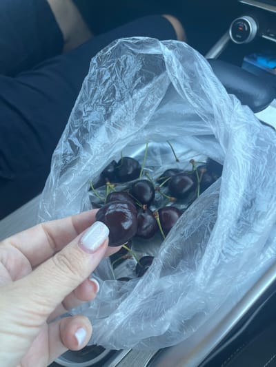 A close-up shot shows a person's hand holding a single dark red cherry, with its stem still attached. The cherry is glistening, suggesting it is fresh. Behind the cherry, a clear plastic bag is filled with more dark red cherries. The bag is partially open, revealing a multitude of cherries inside. The background is blurred but appears to be the interior of a vehicle, with dark seating and a silver console. The scene is lit by natural light, creating a bright and airy feel. There is no indication of the city of Saratovskaya, Russia.