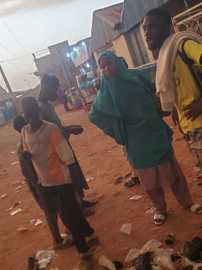 In Funtua, Nigeria, a group of people are gathered on a dusty, unpaved street in front of a row of buildings. The sky is overcast. In the foreground, a young boy in a grey t-shirt with orange and white accents stands next to an adult male wearing dark clothing and a black cap. Behind them, a young girl in a green hijab and pink skirt is talking to a man in a yellow shirt with a tan cloth draped over his shoulder. The man is gesturing with his hands and appears to be speaking. The ground is littered with trash, including plastic bags and wrappers. In the background, a building with a corrugated metal roof is visible, as are shelves stocked with bottled drinks outside a shop. The overall impression is of a bustling street scene in a Nigerian city.