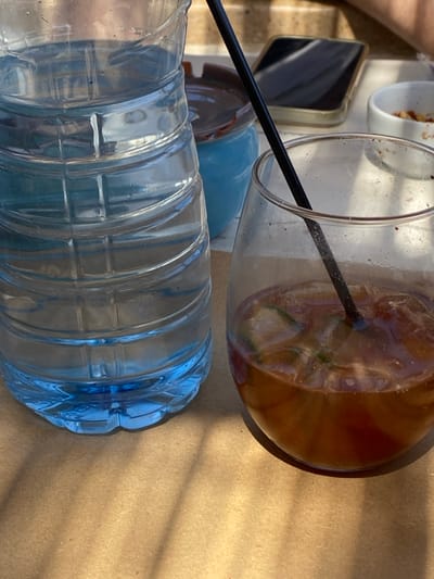 The image displays a close-up, elevated view of a table setting in the city of Φάρος, Greece, illuminated by strong daylight. In the foreground, a clear plastic bottle, approximately half-full with water and exhibiting condensation, stands on the left. Its base is distinctly blue-tinted. To the right, a clear glass tumbler contains a brown, iced beverage, likely a soft drink or cocktail, with several ice cubes and visible green garnishes, possibly lime or cucumber slices. A black plastic straw is inserted into the drink. The glass shows residue along its interior walls.
Behind these two primary objects, a small, light blue ceramic ashtray with a brown rim is visible. Further back and to the right of the ashtray, a dark-colored smartphone with a light-colored or clear case lies flat on the surface. A small, white circular bowl containing a few brown, irregular-shaped items, consistent with nuts or snacks, is partially visible on the far right.
All objects rest on a light brown, flat surface, which appears to be a table or placemat. Strong sunlight from the upper left casts sharp shadows of the bottle, glass, and straw onto this surface, creating distinct patterns. The background shows a blurry, light-colored, textured wall or building exterior, suggesting an outdoor or semi-outdoor setting. No individuals are visible, but the arrangement of items indicates recent human presence or activity.