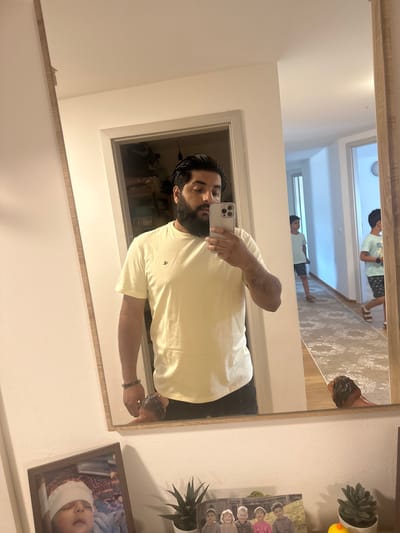 A man with dark hair and a beard is taking a mirror selfie in a hallway. He is wearing a light yellow t-shirt and black pants. In the reflection of the mirror, two young boys are visible walking down the hallway. To the left of the mirror, there are framed photos on a shelf, including one of a baby and another of a group of children. The environment appears to be a residential hallway. The image does not provide specific information about the city of Mainburg, Germany.