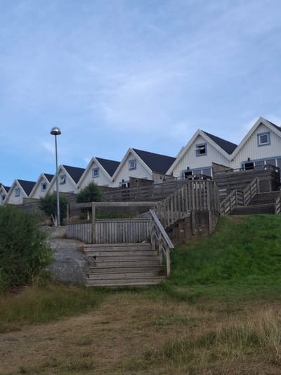 The image depicts a row of identical white wooden houses with dark pitched roofs, situated on a grassy slope in Kebal, Sweden. The houses are arranged linearly, ascending the incline. Each house features triangular gables with a small square window at the apex. Blue trim is visible around the windows and along the roofline. Wooden decks and staircases connect the houses and provide access to them. A tall, slender lamppost with a spherical light fixture stands to the left of the houses. The foreground shows dry grass and some green vegetation. The sky is overcast and a pale blue. There are no people visible in the scene.