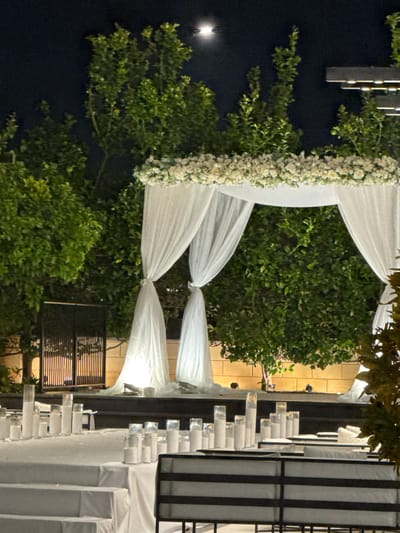 The image depicts an outdoor event setup at night in Ness Ziona, Israel. A white fabric archway, adorned with a dense horizontal arrangement of white flowers across its top beam, occupies the central mid-ground. Sheer white fabric drapes from the archway's corners, gathered and tied back at two points on both the left and right sides. Behind the archway, a dense cluster of dark green trees is visible. The sky above is dark, with a clear, bright moon positioned in the upper center of the frame.

To the far left of the archway, a dark, vertical slatted fence or screen is partially visible. Behind the arch structure, a light-colored wall composed of horizontal blocks is illuminated. In the upper right quadrant, dark, multi-layered architectural elements with integrated lights are visible above the tree line.

In the foreground, multiple tables draped with white tablecloths are arranged on tiered levels. These tables display numerous white pillar candles within clear glass holders of various heights. A seating element with black horizontal slats and white cushions is partially visible in the lower right foreground. No individuals are present. The scene is illuminated by artificial warm lighting, accentuating the white elements, complemented by natural moonlight.
