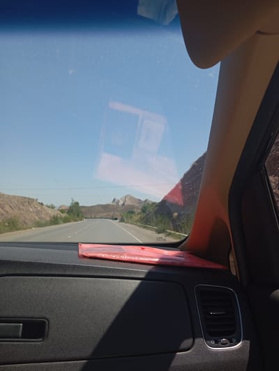 A view from inside a car shows a road stretching through a dry, mountainous landscape under a clear blue sky. The dashboard of the car is visible in the foreground, with a red object, possibly a package or folder, placed on it. The windshield shows reflections, including what appears to be a sun visor. The road is paved and curves slightly to the right, with sparse vegetation and rocky hillsides on either side. The overall impression is of a journey through a remote, arid region. The location is identified as being in the vicinity of Batken, Kyrgyzstan.