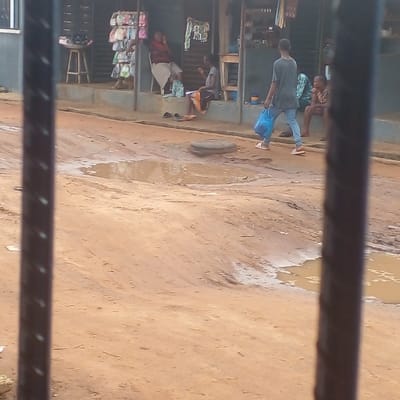 This image depicts a street scene in Ijotan, Nigeria, viewed through two vertical, dark, blurred bars in the immediate foreground. The environment consists of an unpaved, reddish-brown dirt road characterized by numerous ruts, potholes, and puddles of muddy water. A circular, dark-colored manhole cover is positioned in the middle of the road.
On the right side, a single-story building with a light blue-green lower facade features an elevated open porch, indicative of a commercial establishment. Four individuals are present. A young male, wearing a grey t-shirt, light blue jeans, and light-colored shoes, walks from right to left across the dirt road, carrying a blue plastic shopping bag in his left hand. On the building's porch, three individuals are seated. The leftmost seated person, dressed in a reddish top and a light-colored skirt, occupies a chair with their left arm raised. To their right, a second individual, wearing a dark top and an orange-patterned skirt, sits on a lower surface, looking downward. Two pairs of dark shoes are placed on the ground directly in front of this individual. A third seated person, clad in a green and yellow patterned garment, is partially visible on the far right of the porch. Shop merchandise, including a vertical display rack holding multiple colorful bags and other items on a small wooden stool, is visible within the open shop area.