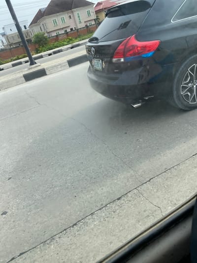 The image depicts the rear passenger side of a dark-colored Toyota Venza SUV, stationary or moving slowly, on a paved road in Igando Oloja, Nigeria. The vehicle exhibits a Nigerian license plate, "LAGOS BOG 951 JN." Light gray exhaust fumes are visibly emitted from the single chrome exhaust pipe located beneath the rear bumper. The rear passenger side tire, featuring a multi-spoke rim, is partially visible. The red taillight assembly includes a distinct blue-colored section.

The road surface is gray asphalt or concrete, showing several cracks. A curb with alternating black and white stripes delineates the roadside to the left of the vehicle. A dark utility pole stands behind this curb.

In the background, a large, two-story light-colored residential building with a reddish-brown hip roof and multiple rectangular windows is prominent, with solar panels visible on its roof. Other residential structures, including one with a red roof, are also visible. Green vegetation and brown perimeter walls separate the road from the background properties. The sky is overcast. A dark, blurred frame element in the bottom right corner suggests the image was taken from inside another vehicle or through a window. No individuals are visible in the scene.
