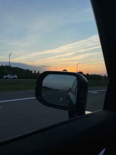The image depicts a view from inside a moving vehicle, looking out towards a sunset. A car's side mirror reflects the road ahead, showing other vehicles and the sky. The exterior shows a highway with a white line marking the lanes, bordered by a grassy area and a dense line of trees in the distance. A lamppost stands on the left side of the road. The sky is filled with wispy clouds, with the sun setting on the horizon, casting a warm orange glow. The car appears to be traveling on a highway in or near the city of Malye Kabany, Russia, given the context provided. There are no people clearly visible in the frame.