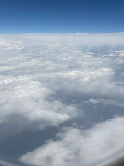 The image displays a view from an airplane window, looking out at a vast expanse of clouds and the sky. On the left side of the frame, a clear, deep blue sky dominates, suggesting good weather and a high altitude. This transitions into a layer of white clouds that fill the majority of the image. The clouds are varied, with some appearing wispy and thin, while others are thicker and more textured, creating a sense of depth and movement. The light suggests it is daytime, likely mid-morning or afternoon, given the brightness and the clear blue of the sky. There are no visible people or specific objects that could indicate a particular location beyond the general setting of being in flight. The overall impression is one of serene vastness, a common experience when flying above the clouds. The image is oriented vertically, so the view is sideways relative to the typical landscape orientation.