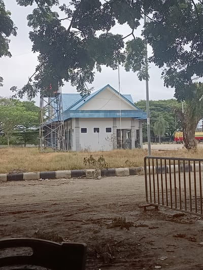 The image depicts an outdoor scene in Meunasah Pupu, Indonesia. In the center of the frame, a white building with a blue roof and trim stands amidst dry, overgrown grass. The building appears to be a small, single-story structure with several windows and a covered entrance. To the left of the building, a metal water tower structure rises, with some scaffolding visible. Trees with dense green foliage are present in the background, partially obscuring the view of a cloudy sky. In the far background, to the right of the building, a yellow and red vehicle, possibly a truck, is partially visible. A metal fence or barrier is in the foreground, and the ground is covered with debris, dirt, and sparse vegetation. The overall impression is one of an unkempt, possibly abandoned or underutilized area.