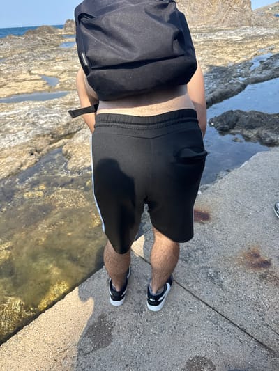 The image shows a person from the waist down, wearing black athletic shorts with a white stripe along the side and black and white sneakers. The person is standing on a concrete path next to a rocky area with shallow water. A black backpack is visible on the person's back, with the word "VANCOUVER" subtly visible on a tag. The lighting suggests it is daytime, with clear skies. The scene appears to be outdoors, possibly near a coast or body of water, with the rocks and water creating a natural environment. The concrete path indicates a developed area within this natural setting. The overall mood is casual and relaxed, consistent with someone engaged in outdoor activity.
