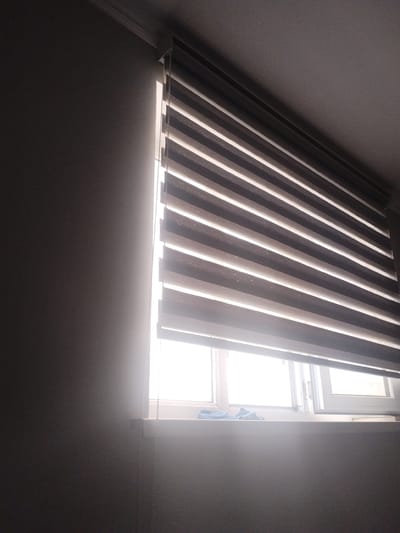 The image shows a window with horizontally slatted blinds partially open, allowing bright daylight to stream in. The blinds are a combination of dark and light colored horizontal stripes. The window is set into a wall which appears to be a muted gray color. The room is dimly lit except for the area illuminated by the sunlight. A light blue cloth or towel is visible on the windowsill. The scene depicts an interior view of a room, likely a home or office, with a window as the central focus. The setting is Vahdat, Tajikistan.