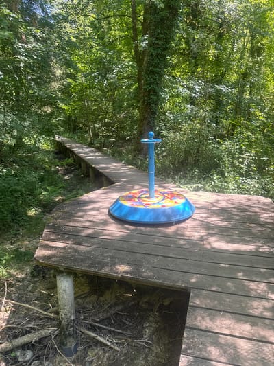 A wooden boardwalk winds through a sun-dappled forest in Ravilloles, France. In the foreground, a circular, blue, outdoor game or play structure with colorful geometric patterns sits atop a wooden platform. The structure has a central blue pole with a knob at the top. The boardwalk is elevated, supported by wooden posts, and appears to be constructed from weathered planks. The surrounding environment is dense with green foliage, including trees and undergrowth, with sunlight filtering through the leaves. A small, dry creek bed or ditch is visible beneath a section of the boardwalk. The scene suggests a recreational area within a natural setting.