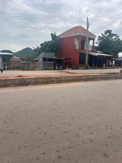 The image shows a street scene in Kona Garu, Nigeria. The scene is dominated by a two-story red building with balconies and windows. In front of the building are some smaller structures, possibly shops or shelters, made of corrugated metal and wood. There are a few people visible in the background, some tending to market stalls. The street itself is paved and has a distinct curb with rough texture. The sky is overcast, suggesting a cloudy day. In the distance, a lush green hill or mountain range is visible, adding a natural element to the otherwise urban landscape. Power lines run across the scene, indicating the presence of electricity infrastructure. The overall impression is of a busy, everyday scene in a Nigerian town, possibly during market hours.